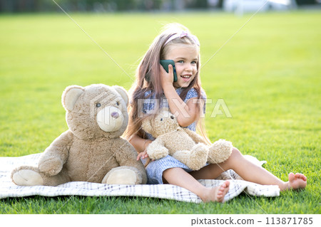 Pretty child girl sitting in summer park on green grass lawn with her teddy bear toy talking on mobile phone outdoors in summer. Pretty child girl sitting in summer park on green grass lawn with her teddy bear toy talking on mobile phone outdoors in summer. 113871785