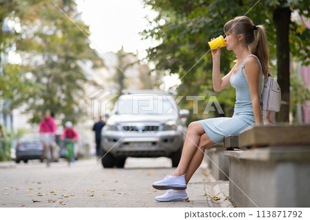 Young woman drinking coffee from paper cup sitting on city street bench in summer park. 113871792