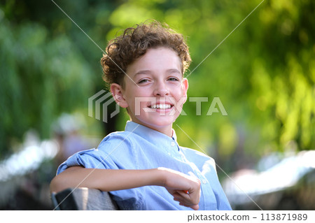 Young happy child boy relaxing sitting on bench in summer park. Positive kid enjoying summertime outdoors. Child wellbeing concept Young happy child boy relaxing sitting on bench in summer park. Positive kid enjoying summertime outdoors. Child wellbeing concept 113871989