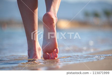 Close up of female feet walking barefoot on white grainy sand of golden beach on blue ocean water background 113871999
