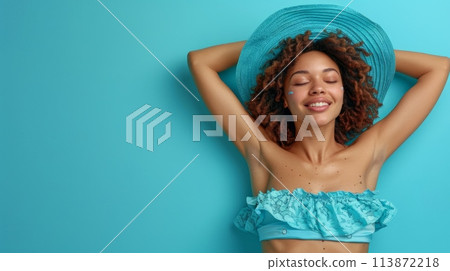 A woman with flowing hair stands on a beach in a blue bikini top and hat, gazing out at the sparkling sea under the bright sun. 113872218