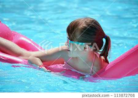 Happy child girl relaxing on inflatable air mattress in swimming pool on sunny summer day during tropical vacations. Summertime activities concept 113872708