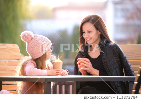Young pretty woman having good time with her child daughter sitting at street cafe with hot drinks on sunny autumn day. Happiness in family relations concept 113872784