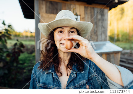 Funny Woman Farmer in denim with a fresh egg, making a funny face. A woman in hat exhibits sheer happiness with a fresh egg in hand, standing on her farm Sustainable lifestyle, organic farmer Funny Woman Farmer in denim with a fresh egg, making a funny face. A woman in hat exhibits sheer happiness with a fresh egg in hand, standing on her farm Sustainable lifestyle, organic farmer 113873744