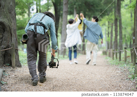A cameraman uses a gimbal to film a family walking through the fresh greenery A cameraman uses a gimbal to film a family walking through the fresh greenery 113874047