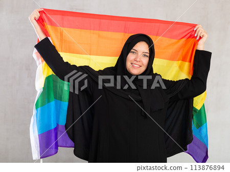 Happy young woman dressed in traditional islamic clothes with rainbow flag 113876894