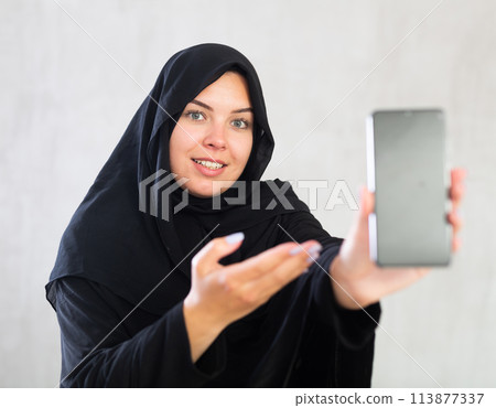 smiling Muslim woman in traditional black hijab shows blank smartphone screen close-up smiling Muslim woman in traditional black hijab shows blank smartphone screen close-up 113877337