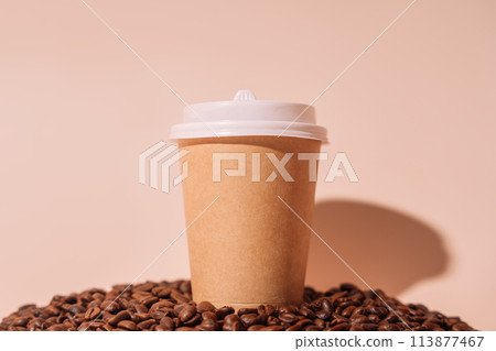 Paper cup with takeaway coffee on a pile of coffee beans against a beige background 113877467