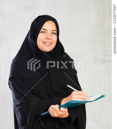 gleeful girl in hijab holds several notebooks and writes important note in her diary gleeful girl in hijab holds several notebooks and writes important note in her diary 113877487