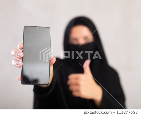 young Muslim girl with her face covered by burka shows empty dark smartphone screen. young Muslim girl with her face covered by burka shows empty dark smartphone screen. 113877558