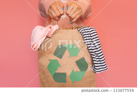 Paper bag with recycling sign with old clothes in hands against pink background Paper bag with recycling sign with old clothes in hands against pink background 113877559
