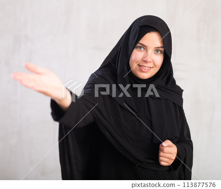 Cheerful young muslim woman dressed in black hijab pointing at side Cheerful young muslim woman dressed in black hijab pointing at side 113877576