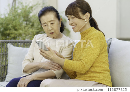A senior woman and a middle-aged parent and child sitting on a sofa and talking while looking at a smartphone 113877825