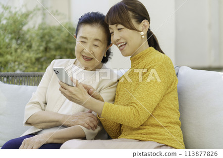 一位老年婦女和一位中年父母和孩子坐在沙發上，一邊看著手機一邊聊天。 113877826