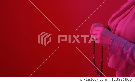 Wooden rosary. Ramadan pray. Unrecognizable woman praying with tasbih beads in hands in neon light on red copy space background. 113885900