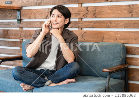 Young Asian man sitting on a sofa at home talking on his smartphone Young Asian man sitting on a sofa at home talking on his smartphone 113886368