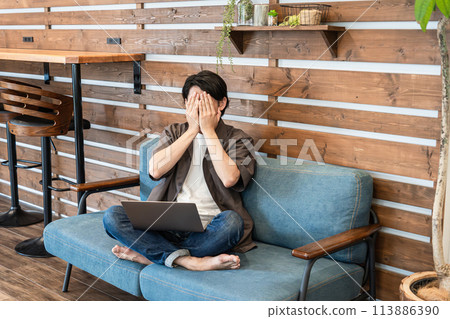 Young man covering his face while looking at a laptop on the sofa at home (troubled, regretful, miserable, shameful, terrible, cruel, trouble) Young man covering his face while looking at a laptop on the sofa at home (troubled, regretful, miserable, shameful, terrible, cruel, trouble) 113886390