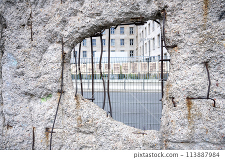 This photograph captures a close-up view of a hole in a remaining section of the Berlin Wall. The weathered texture and exposed, rusted rebar tell a story of age and exposure to elements. Through the 113887984