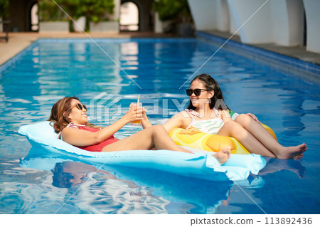 Smiling mother and daughter spending time in swimming pool chatting and drinking cocktails 113892436
