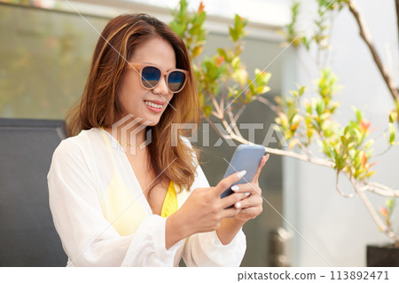 Portrait of smiling woman in beach robe and sunglasses checking messages on smartphone Portrait of smiling woman in beach robe and sunglasses checking messages on smartphone 113892471