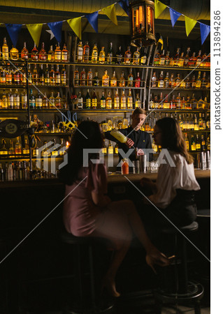 Barkeeper pours syrup into jigger standing near female guests Barkeeper pours syrup into jigger standing near female guests 113894286