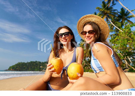 Two friends enjoy beach vacation, sitting on sand with fresh coconuts, wearing sunglasses and swimwear, posing at tropical palm-fringed shore, sunny blue sky reflects a perfect holiday retreat. 113900833