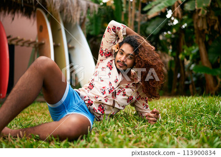 Confident gay man with rich curly hair reclines in tropical garden, vibrant style. Blue shorts, floral shirt enhance vivid look. LGBT travel, self-love ambiance resonate. Leisure in natural, sunny. Confident gay man with rich curly hair reclines in tropical garden, vibrant style. Blue shorts, floral shirt enhance vivid look. LGBT travel, self-love ambiance resonate. Leisure in natural, sunny. 113900834