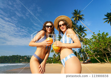 Happy women in bikinis enjoy fresh coconuts on sunny beach. Friends share exotic vacation by blue sea, green palms. Travel lifestyle, leisure relax, nature adventure. Girls sunshine holiday. 113900838