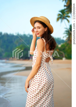 Smiling woman in polka dot dress and straw hat walks on sunny beach. Retro summer fashion, relaxed atmosphere. Palm trees, blue sky in background. Female enjoys seaside, warm weather, coastal vibes. 113900844