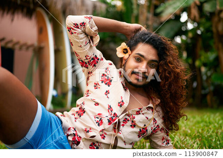Gay man with floral shirt poses, holds flower, exudes confidence, style in rich garden. Vibrant outdoor setting for travel, lifestyle blogs. LGBT-friendly tropical vacation scene. 113900847