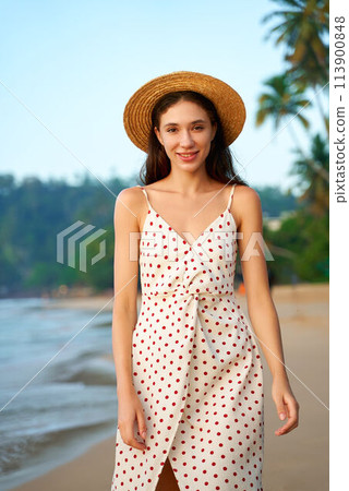 Woman in polka dot dress and straw hat smiles on sunny beach. Retro summer fashion on display. Female enjoys seaside, tropical backdrop with blue sky, soft waves. Holiday scene, relaxed vibe. Woman in polka dot dress and straw hat smiles on sunny beach. Retro summer fashion on display. Female enjoys seaside, tropical backdrop with blue sky, soft waves. Holiday scene, relaxed vibe. 113900848