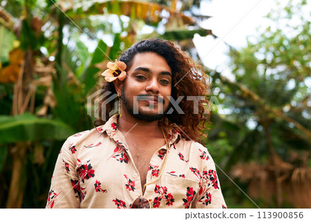 Confident man with flower in hair poses in nature. Gay, showing pride, diversity, radiates happiness. Outdoors, represents inclusion, nature beauty. Floral shirt complements tropical setting. Confident man with flower in hair poses in nature. Gay, showing pride, diversity, radiates happiness. Outdoors, represents inclusion, nature beauty. Floral shirt complements tropical setting. 113900856