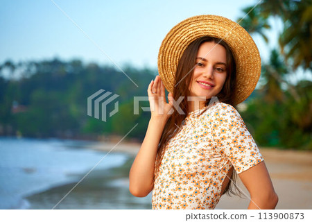 Smiling woman in floral shirt, straw hat stands on tropical beach at sunset. Relaxed female enjoys summer evening by sea, sandy shore, vacation vibe. Retro beach fashion, leisure concept. Smiling woman in floral shirt, straw hat stands on tropical beach at sunset. Relaxed female enjoys summer evening by sea, sandy shore, vacation vibe. Retro beach fashion, leisure concept. 113900873