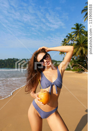 Happy woman in bikini holds fresh coconut on sunny tropical beach. Female enjoys summer by sea with palm trees. Blue sky, clear ocean water, wellness lifestyle. Drinking natural refreshment. Happy woman in bikini holds fresh coconut on sunny tropical beach. Female enjoys summer by sea with palm trees. Blue sky, clear ocean water, wellness lifestyle. Drinking natural refreshment. 113900888