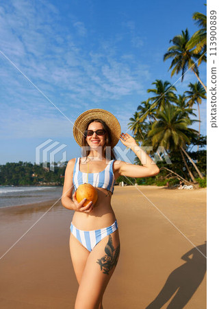 Woman in striped bikini holds fresh coconut on sunny beach. Solo traveler enjoys tropical seaside, healthy drink. Palm trees, blue sky in background. Relaxing, hydration benefits by the sea. 113900889
