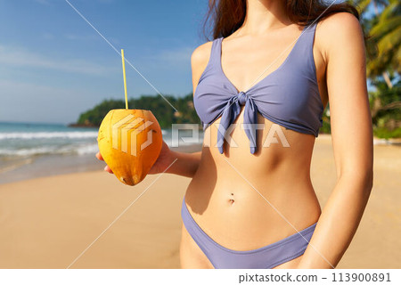 Close-up of fit woman in bikini holding fresh coconut with straw on tropical beach. Sunny travel destination, healthy lifestyle, thirst quenching, summertime leisure by the sea. 113900891