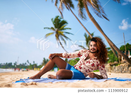 Gay man relaxes on beach, enjoys sunbath, lying on towel under palms. Sunny day, clear sky in tropical paradise during Pride Month. Contentment, leisure on sandy shore, feeling free and peaceful. Gay man relaxes on beach, enjoys sunbath, lying on towel under palms. Sunny day, clear sky in tropical paradise during Pride Month. Contentment, leisure on sandy shore, feeling free and peaceful. 113900912