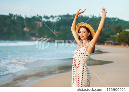 Happy woman in straw hat, polka dot dress raises arms on sandy beach. Smiling female enjoys tropical sun, waves, seaside vacation. Solo traveler explores coastal landscape, freedom concept. 113900921