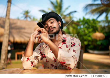 Gay man relaxes at beach cafe, palm trees sway. Stylish male enjoys tropical getaway, sits at wooden table. Casual, confident look, exotic vacation, LGBTQ travel destination, sunny seaside retreat. 113900922