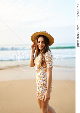 Smiling woman in vintage floral dress and straw hat walks on sandy beach. Blue ocean waves in background. Summer, fashion, leisure concept. Happy female enjoys sunset by the sea, retro style. Smiling woman in vintage floral dress and straw hat walks on sandy beach. Blue ocean waves in background. Summer, fashion, leisure concept. Happy female enjoys sunset by the sea, retro style. 113900937