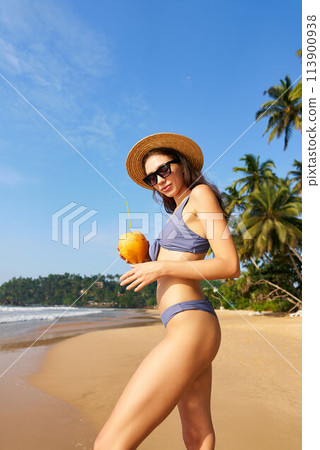 Smiling fit woman in sunglasses and straw hat holds fresh coconut on sunny tropical beach. Bikini-clad traveler enjoys healthy drink, palm trees, blue sky background. Wellness, summer holiday scene. 113900938