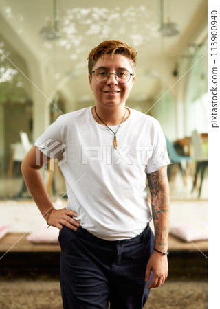 Smiling Gen Z transgender pro stands in modern office, casual style wear, tattoo visible, hands in pockets, gender diversity at work, embodying inclusive corporate culture, workplace equality. Smiling Gen Z transgender pro stands in modern office, casual style wear, tattoo visible, hands in pockets, gender diversity at work, embodying inclusive corporate culture, workplace equality. 113900940