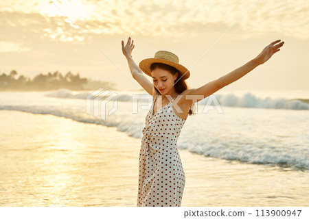 Elegant lady in polka dot dress enjoys sunset at sea beach, arms raised in joy. Solo traveler celebrates freedom, summertime bliss on tropical coast. Happiness, leisure, ocean breeze felt by tourist. 113900947