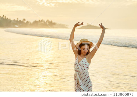 Woman in polka dot dress enjoys sunset at beach, arms raised high. Solo traveler celebrates freedom, warm summer evening by sea. Happiness, relaxation with ocean waves, sandy shore in background. 113900975