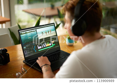Pro video editor with headphones works on laptop in cafe. Over shoulder view, editing software screen, colorful timeline. Camera, glasses on table. Freelancer edits travel vlog, ambient background. Pro video editor with headphones works on laptop in cafe. Over shoulder view, editing software screen, colorful timeline. Camera, glasses on table. Freelancer edits travel vlog, ambient background. 113900989