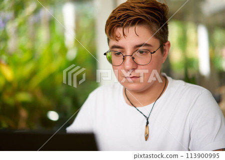 Transgender pro focuses laptop work in office. Inclusive environment, modern workplace diversity. Confident individual with glasses, casual attire. Background plants indicate eco-friendly space. 113900995