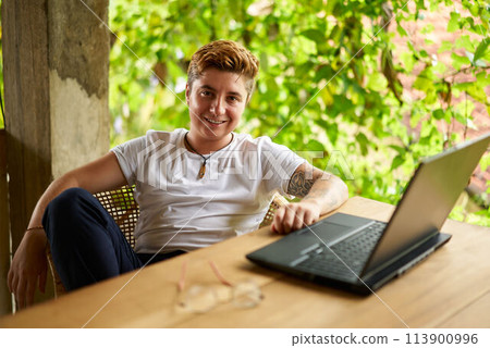 Transgender Gen Z with tattoos smiles, works on laptop in tropical coworking space. Inclusive remote work scene captures gender diversity, modern freelancing trend amongst rich plants. Transgender Gen Z with tattoos smiles, works on laptop in tropical coworking space. Inclusive remote work scene captures gender diversity, modern freelancing trend amongst rich plants. 113900996