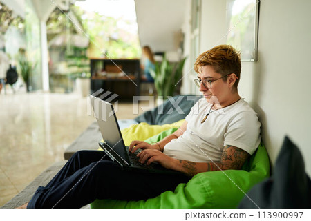 Transgender professional works on laptop, seated on green bean bag, in casual office space. Relaxed environment, light-filled room, modern work life balance, inclusion in workplace highlighted. 113900997