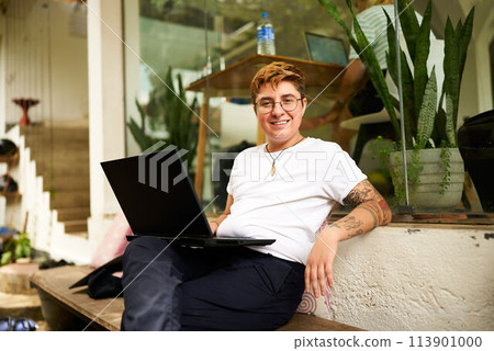 Transgender Gen Z freelancer smiles while working on laptop at eco-friendly coworking space. Inked, digital nomad in casual clothes, emphasizing diversity, inclusivity in modern remote work settings. Transgender Gen Z freelancer smiles while working on laptop at eco-friendly coworking space. Inked, digital nomad in casual clothes, emphasizing diversity, inclusivity in modern remote work settings. 113901000