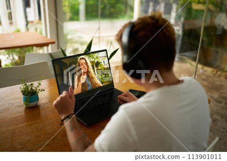 Gen Z pro in casual office setup participates in video call from home. Young person with headphones communicates online, discusses project with colleague. Modern work from anywhere concept. 113901011